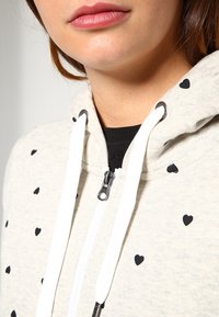 Sudadera con capucha gris claro con cremallera y pequeños patrones de corazones negros. Cuenta con cordones blancos y herrajes metálicos. Tejido suave con un ajuste relajado.