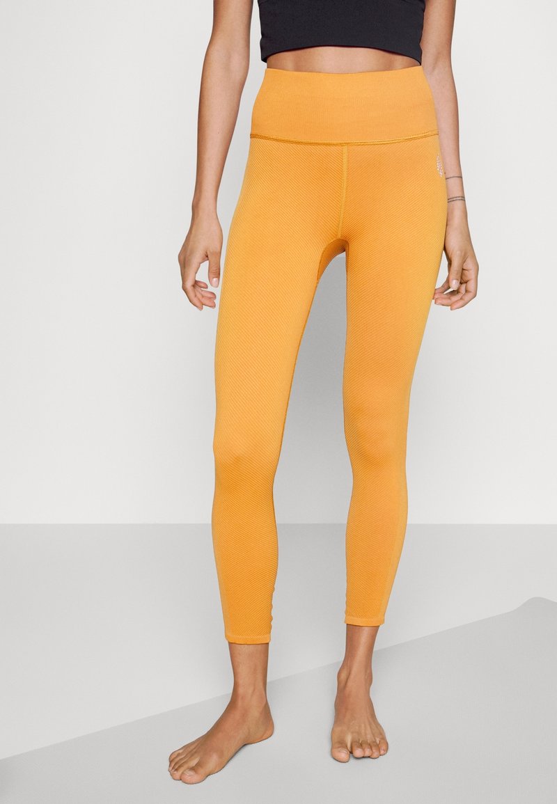 Mujer vestida con leggings naranja de alta cintura y textura, de pie descalza sobre un suelo gris bicolor con los brazos relajados a los lados.