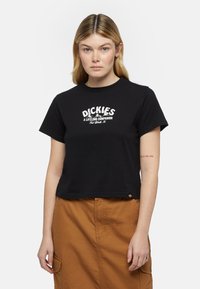 Černé zkrácené tričko s bílým nápisem "DICKIES A LIFELONG COMPANION". Má krátké rukávy a rovný lem. Hnědá sukně je viditelná dole.