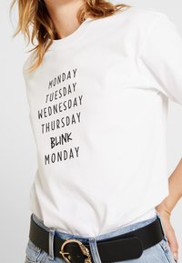 Bílá bavlněná trička s černým textem vyjmenovávajícím dny v týdnu a "BLINK MONDAY". Kulatý výstřih, krátké rukávy, volný střih.