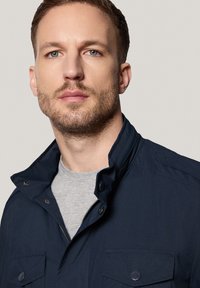 Granatowa kurtka z stojącym kołnierzem, zapinana na zamek, z zatrzaskami. Posiada dwie kieszenie na piersiach i gładką teksturę. Pod spodem widoczna szara koszulka.