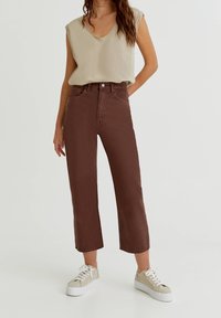 Jean marron taille haute avec une coupe courte et jambes droites. Associé à un haut sans manches beige clair et des baskets blanches.