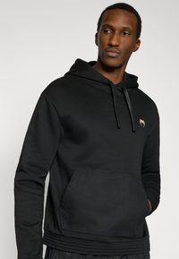 Μαύρο φούτερ με κουκούλα και υφασμένη μπροστινή τσέπη· διαθέτει μικρό λογότυπο στο στήθος και ριμπ λεπτομέρειες στο τελείωμα και στα μανίκια.