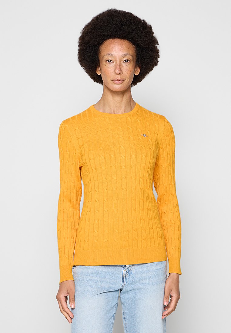Żółty sweterek z dzianiny o teksturowanym wzorze w warkocze i okrągłym dekolcie. Posiada długie rękawy i mały detal z logo na klatce piersiowej.