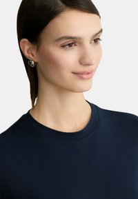 Camicia in cotone blu navy con scollatura rotonda, caratterizzata da una texture liscia. Orecchini a forma di cuore argentati aggiungono un accento sottile.