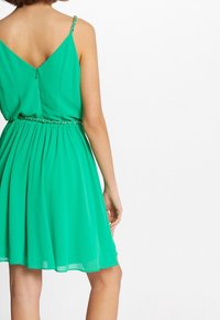 Grön chiffongklänning med V-rygg, tunna kedjespännen, veckad kjol och en dekorativ bälte detalj i midjan. Slät textur genom hela.