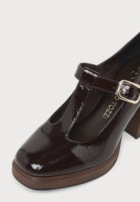Chaussure Mary Jane en cuir verni marron foncé avec bride en T, boucle dorée, bout arrondi et talon épais en bois.