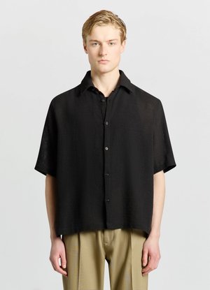 Young man with blond hair wearing a loose black short-sleeve button-up shirt and beige pants against a plain light background.