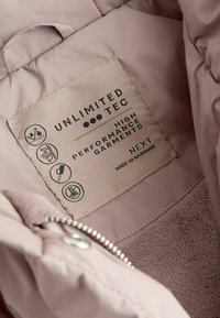 Étiquette en tissu rose cousue dans un vêtement, présentant des symboles d'entretien, "Unlimited TEC" et "Next", indiquant un matériau haute performance.