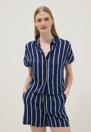 Jeune femme aux cheveux blonds et raides porte une chemise bleu marine à manches courtes boutonnée et un short à rayures assorti, mains dans les poches.