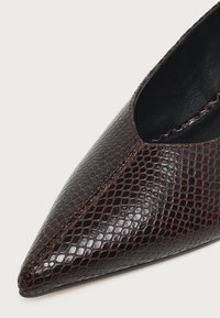 Gros plan d'une chaussure à bout pointu marron foncé avec un motif texturé rappelant la peau de serpent et des coutures visibles le long des bords.