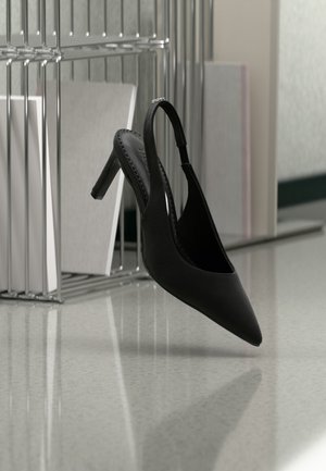 Čierna vysoká podpätková obuv s otvorenou pätou a špicatou špičkou, visiaca vo vzduchu nad lesklou podlahou, v pozadí sú stohované šanóny.