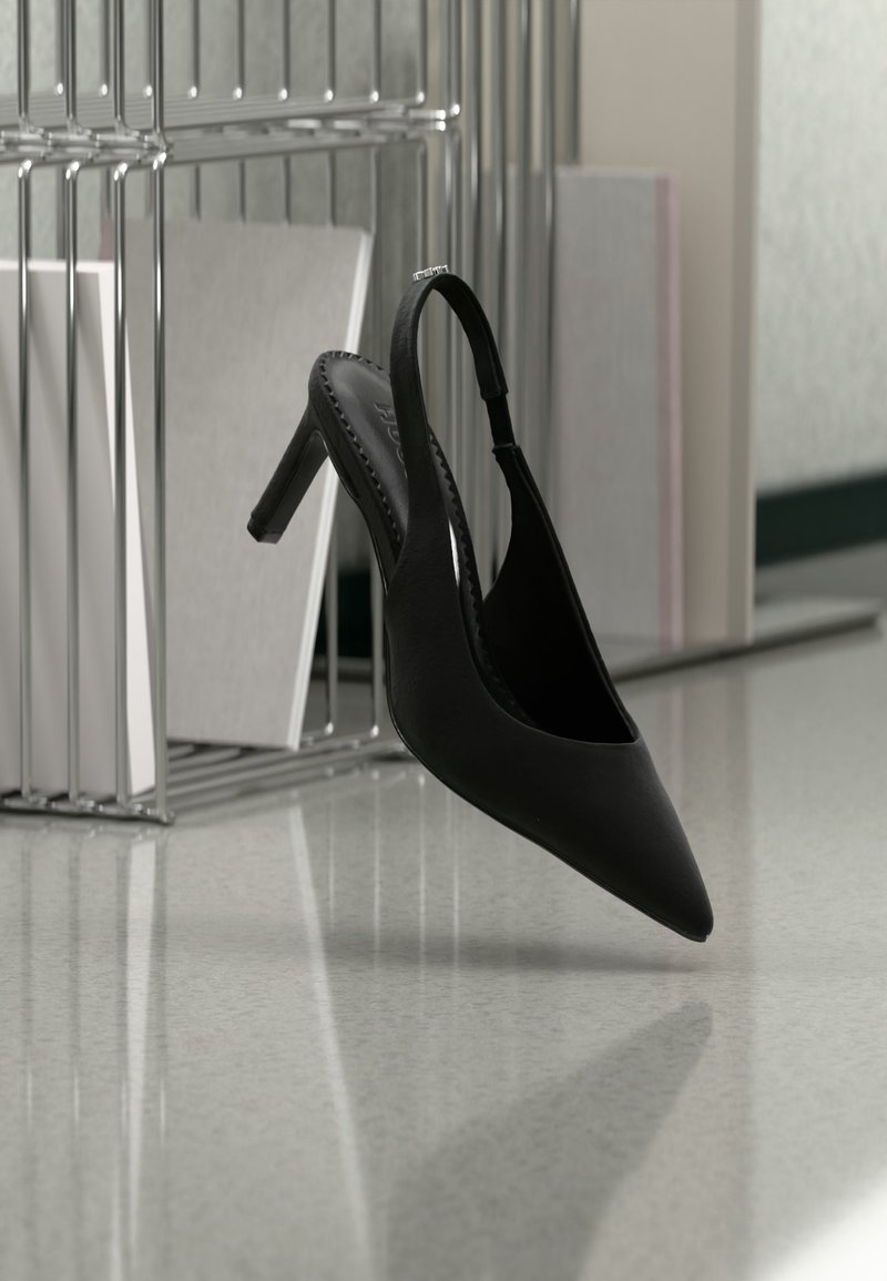 Μαύρο ψηλοτάκουνο παπούτσι με μυτερή μύτη και λουράκι πίσω, κρεμασμένο στον αέρα πάνω από ένα γυαλιστερό πάτωμα με στοιβαγμένους φακέλους στο φόντο.