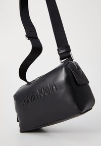 Černá kožená crossbody taška s texturovaným povrchem, nastavitelným popruhem a stříbrným zipem. Obsahuje vyražené logo "Calvin Klein".
