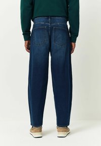 Sötétkék, lazán illeszkedő farmernadrág, két hátsó zsebbel és klasszikus derékpánttal, bézs sportcipővel párosítva.