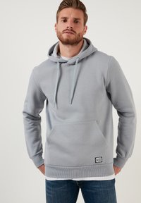 Sudadera de algodón gris con un bolsillo frontal, drawstrings ajustables, puños acanalados y una etiqueta de la marca cerca del dobladillo. Textura suave.