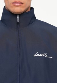 Jūros laivyno vėjui atsparus švarkas su aukštu apykakle, visišku užtrauktuku ir baltu "Lacoste" logotipu. Lygus paviršius, minimalistiškas dizainas ir patvari medžiaga.