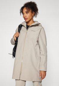 Beige vanntett jakke med glidelås foran, justerbar hette og sidelommer. Glatt tekstur og avslappet passform, designet for utendørsbruk.