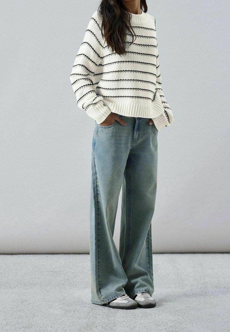 Femme portant un pull en maille blanc à rayures noires, un jean ample bleu clair et des baskets grises, debout, les mains dans les poches.