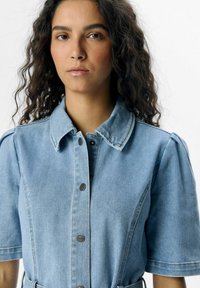 Vestido de camisa de denim azul claro que presenta un frente abotonado, mangas cortas, cuello puntiagudo y una textura sutil con una silueta ajustada.