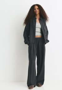 Femme aux longs cheveux bouclés portant un débardeur gris, une chemise noire oversize à boutons, un pantalon noir ample et des baskets, se tenant contre un mur blanc.