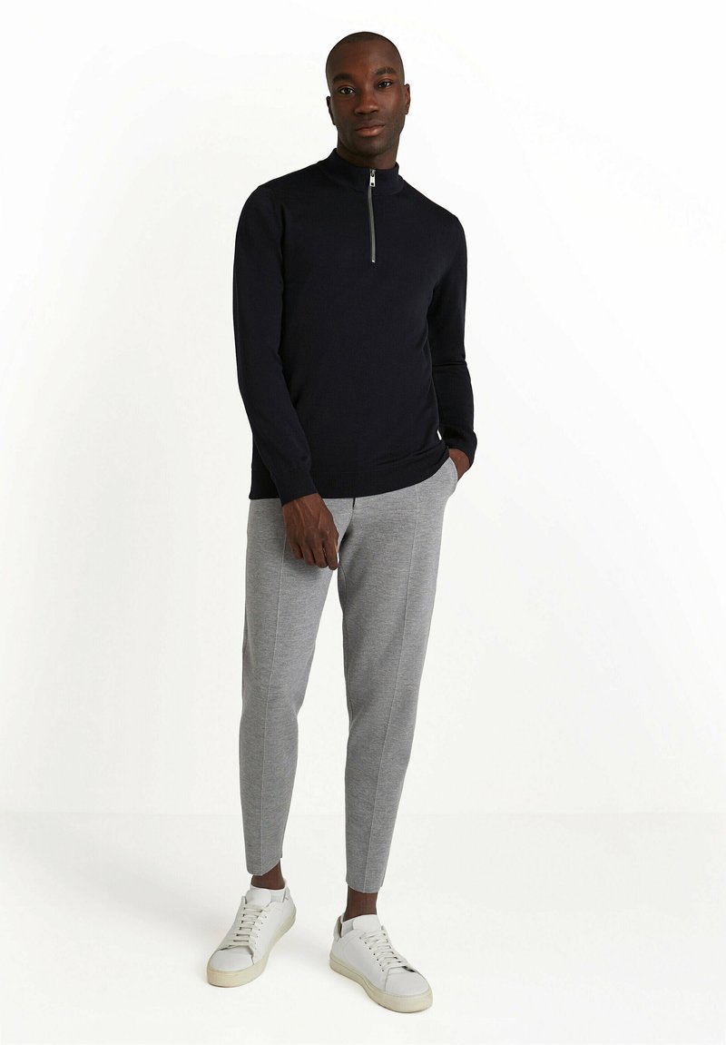 Maglione navy a zip e maniche lunghe, pantaloni jogging aderenti grigi chiari e sneakers bianche con finitura testurizzata e lacci visibili.