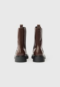 Pár vysokých hnědých kožených bot s černými masivními podrážkami, zobrazených zezadu na jednobarevném světlém pozadí.