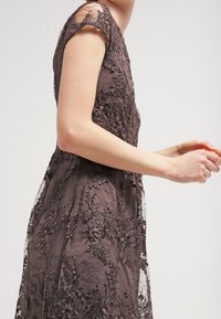 Robe en dentelle marron foncé avec broderies florales, manches courtes et corsage ajusté, présentant une superposition transparente et des détails complexes.