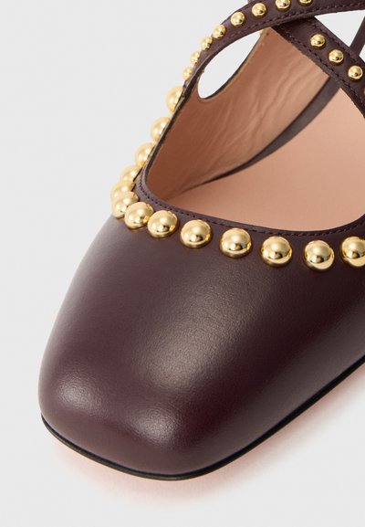 Scarpa in pelle borgogna con punta quadrata, caratterizzata da una fila di borchie metalliche dorate lungo il bordo e un design a fascette incrociate.