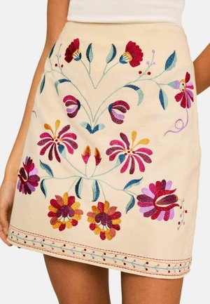 Beige Rock mit bunten, gestickten Blumen in Rot, Lila, Gelb und grünen Blättern, getragen mit einem weißen Oberteil.