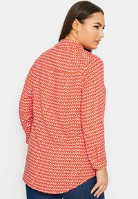 Blusa rossa con motivo a diamante bianco, maniche lunghe, vestibilità rilassata, dettaglio di piega sul retro e orlo arrotondato. Realizzata in tessuto leggero.