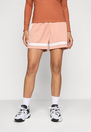 Person trägt pfirsichfarbene Sportshorts mit weißen Streifen, weiße Socken, weiße und schwarze Sportschuhe und ein langärmeliges Oberteil in Rostfarbe.