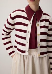 Pruhovaný cardigan v krémové a burgundské barvě, s texturovaným pletením, zlatými knoflíky a kulatým límcem, nošený přes burgundový top a béžovou sukni.