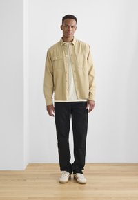 Beige kordfløyels skjortebluse med to brystlommer, lagd over en kremfarget t-skjorte, kombinert med svarte bukser og hvite sneakers.