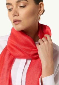 Cienki, lekki czerwony szalik zwisający na szyi, o gładkiej fakturze, noszony na białym ubraniu. Tkanina wydaje się miękka i delikatna.