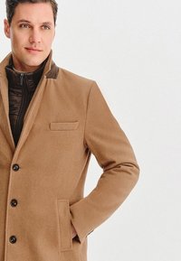 Brązowy płaszcz z mieszanki wełny z klapkowym kołnierzem, zapięciem na guziki i bocznymi kieszeniami. Posiada kontrastowy ciemny materiał przy kołnierzu i zapinaną na zamek warstwę wewnętrzną.