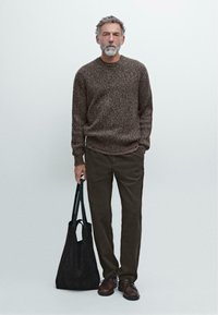 Brun tekstureret striktrøje, mørkebrun løstsiddende bukser, mørk ruskinds tote bag og brune sko. Modellen står foran en ensfarvet baggrund.