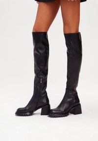 Botas negras por encima de la rodilla hechas de suave cuero sintético, con un tacón grueso, suela texturizada y cremallera lateral para facilitar el uso.
