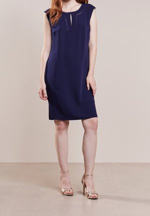 Femme portant une robe sans manches bleu marine jusqu'aux genoux avec un détail de décolleté rectangulaire et des sandales à talons hauts dorées.