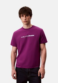 T-shirt violet en coton avec col rond et manches courtes, arborant un logo blanc imprimé sur le devant. Le tissu semble lisse et léger.