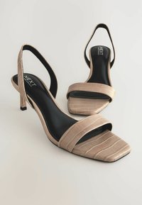 Sandales à talons bas en simili-crocodile beige, à bout carré, avec de larges brides arrière. Semelle intérieure noire visible.