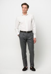 Camicia bianca a maniche lunghe con bottoni, infilata in pantaloni grigi a pieghe, abbinata a mocassini marroni scuri. L'outfit ha un aspetto classico e sartoriale.