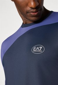 Camiseta atlética azul-marinho com tecido texturizado, apresentando um detalhe roxo e o logótipo EA7 no peito. Mangas curtas e decote redondo.