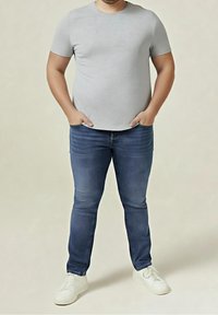 Man standing with hands in pockets wearing a light grey t-shirt, blue jeans, and white sneakers on a neutral background.