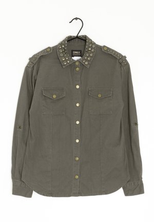 Olivově zelená košile s hroty na límečku, dvěma předními kapsami a mosaznými knoflíky. Dlouhé rukávy s možností ohrnutí a zaoblený lem.