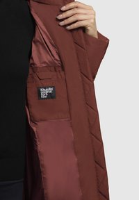 Ranka laiko atvirą bordo spalvos siuvinėta striukę, rodydama vidinį kišenę su juodu etiketu, kuriame parašyta "Khujo original for a few."