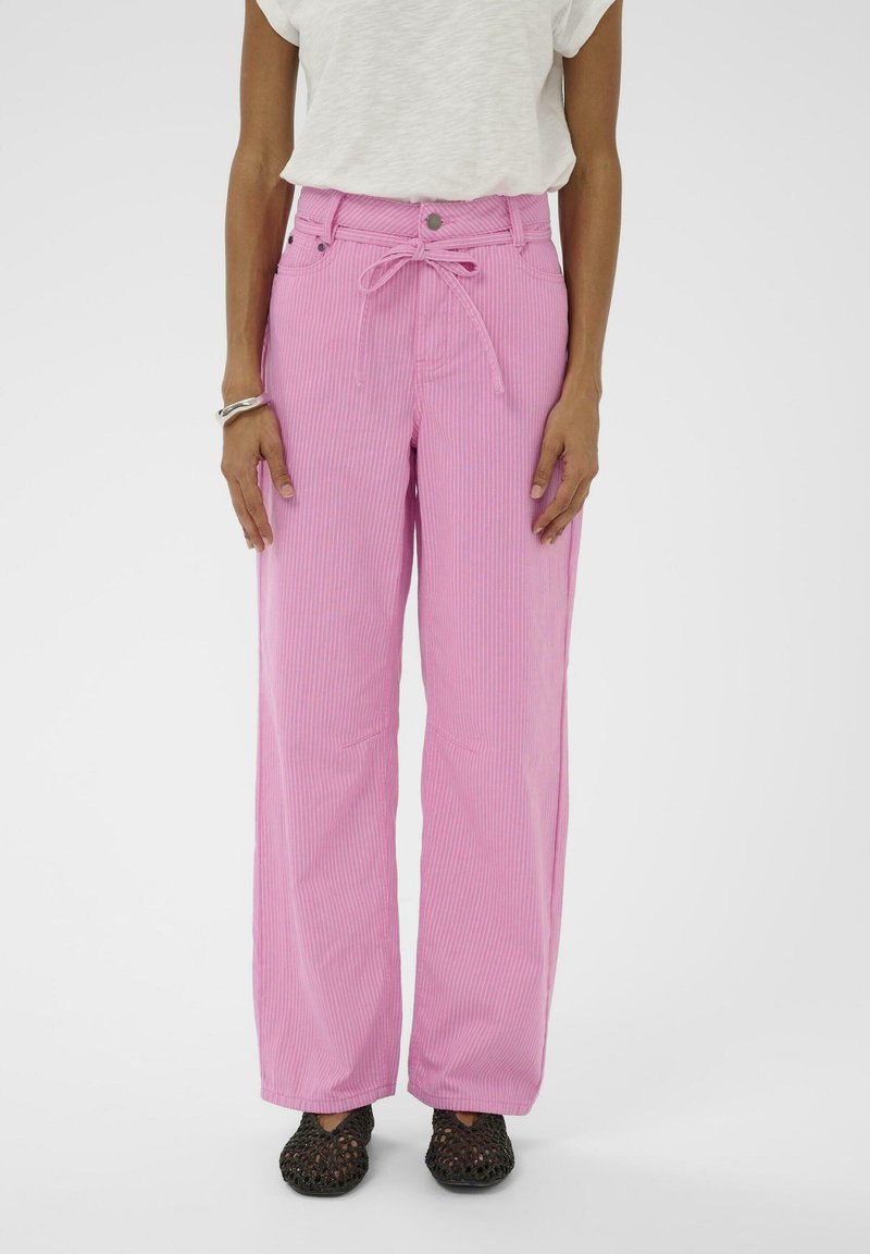 Személy magas derekú rózsaszín kordbársony nadrágban, megkötött övvel, fehér rövid ujjú felsőben, ezüst karkötővel és fekete szövött cipőben.