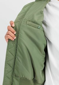 Interiorul jachetei matlasate de coloră verde măslin, textura mătăsoasă, are un buzunar lateral cu un buton tip snap. Țesătură cu nervuri la tiv și la mansete, cu branding discret.