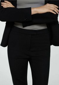 Blazer noir sur mesure avec une texture subtile, doté d'une fermeture à un bouton unique et d'un pantalon noir slim avec une finition lisse. Haut gris en dessous.