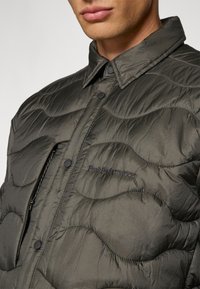 Chaqueta acolchada verde oscuro con un collar abotonado, que cuenta con un bolsillo con cremallera y un sutil logo bordado en el pecho. Tejido texturizado.